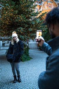 Cropped image of man photographing girlfriend standing against christmas tree in city