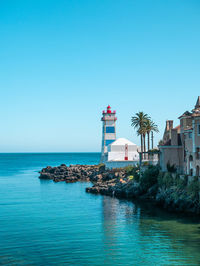 Lighthouse by sea against buildings against clear blue sky