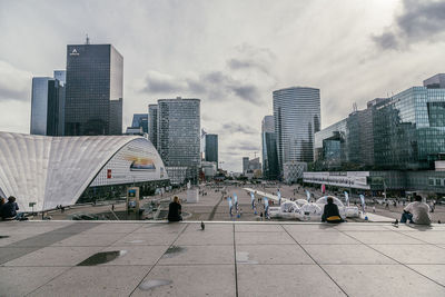 People on footpath amidst modern buildings against cloudy sky