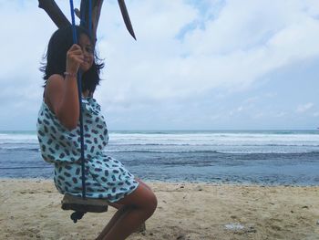 Side view of girl sitting on swing at beach against sky