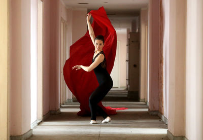 Full length portrait of young woman doing ballet dance with red fabric in corridor