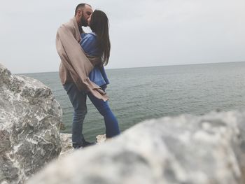 Side view of couple kissing on rock by sea against sky