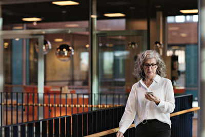 Female senior professional with headphones using smart phone while walking in atrium at office