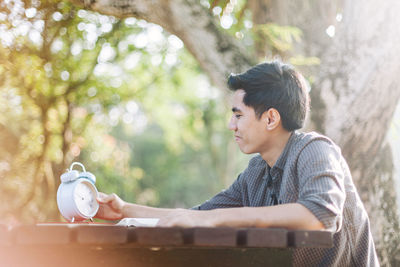 Young man looking at alarm clock while sitting at table in park