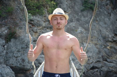 Portrait of shirtless man gesturing thumbs up on bridge against rock formation