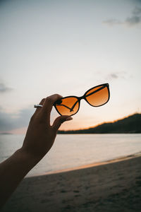 Woman holding sunglasses on beach against sky during sunset