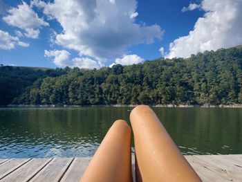 Woman with her tanned legs resting on a wooden pontoon near a lake surrounded by forest