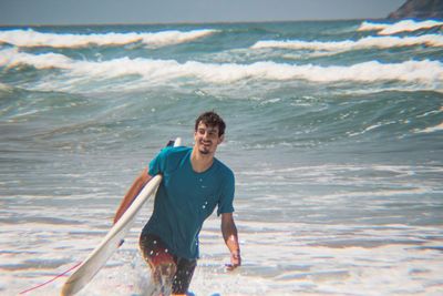 Smiling young man holding surfboard while walking in sea