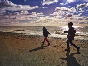 Side view of brothers walking at beach against cloudy sky during sunset