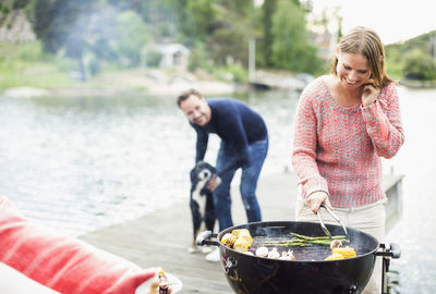 Happy woman using mobile phone while barbecuing with man and dog in background on pier