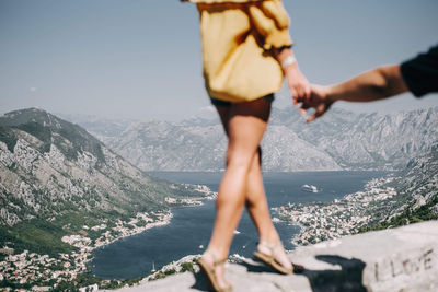 Low section of woman holding man hand while walking on retaining wall against mountain