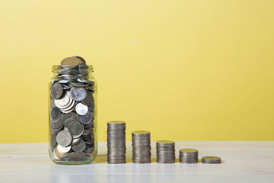 Coins and jar against yellow background on table