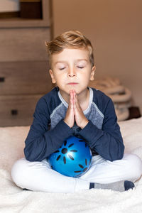 Cute boy praying while sitting on bed at home