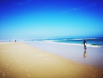 Rear view of surfer standing on shore at beach against blue sky