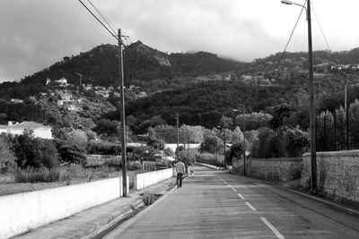 Rear view of man walking on road by mountain against cloudy sky