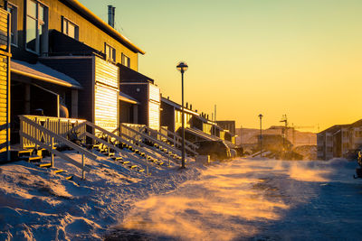 Snow covered street by buildings against sky during sunset