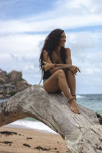 Woman sitting on log at beach against sky