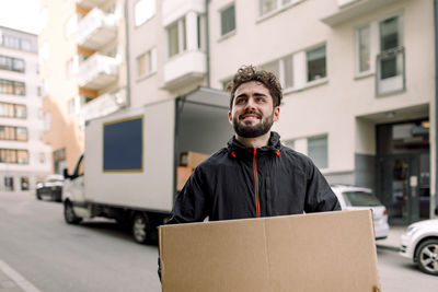 Confident young delivery man carrying cardboard box in city