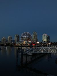 Illuminated modern buildings by river against clear sky