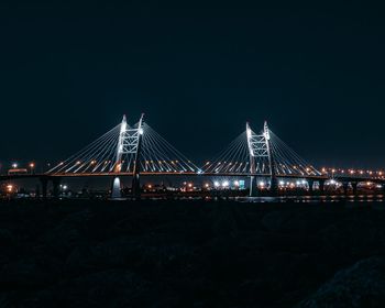 Illuminated suspension bridge over river against sky at night