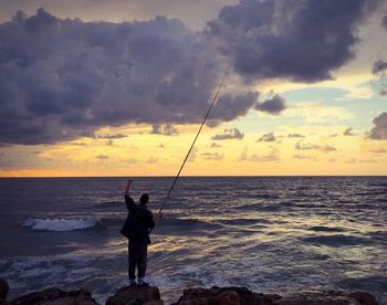 Silhouette man fishing in sea against sunset sky