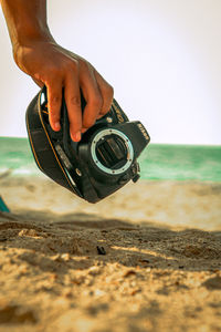 Hand holding camera at beach against clear sky