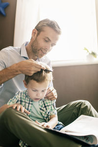 Boy reading book while father combing his hair at home