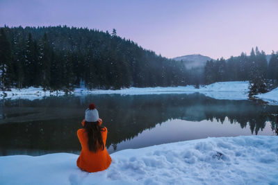 Rear view of woman sitting by lake during winter