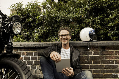 Laughing man with tablet sitting on the ground beside his motorbike