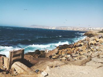 Scenic view of splashing sea waves on rock against clear blue sky