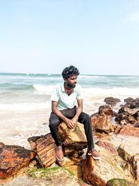 Young man sitting on rock by sea against sky