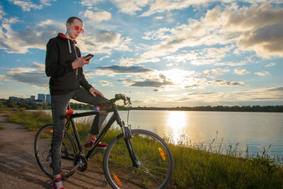 Man with bicycle standing by lake against sky during sunset