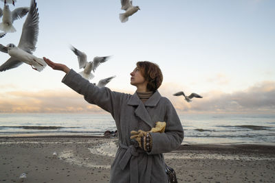 Young woman in overcoat feeding seagulls at beach