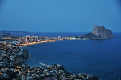 Aerial view of illuminated city by sea against clear blue sky