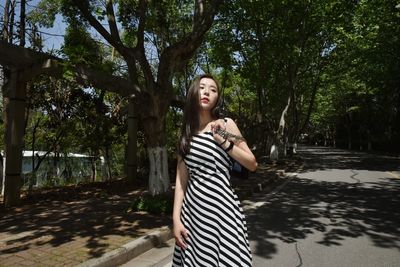 Young woman looking away while standing on road against trees during sunny day