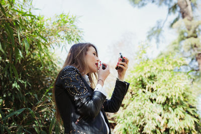 Low angle view of mid adult woman applying lipstick while standing against plants in park