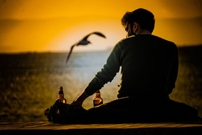 Rear view of man drinking while sitting at beach during sunset