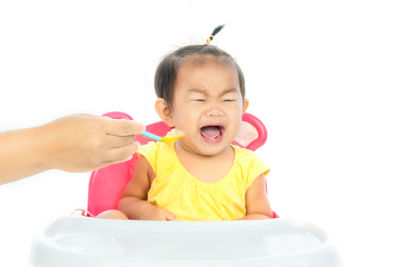 Cropped hand of mother feeding crying daughter sitting on high chair against white background