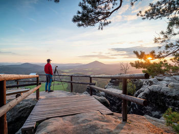 Hotographer in red hoodie stay at camera on basalt tripod on rocky cliff with wooden handrail around