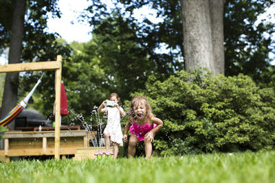 Girl photographing while sister playing with sprinkler in backyard