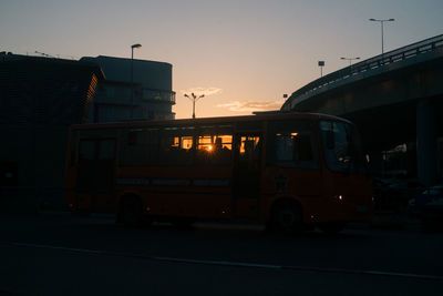 Cars on road by buildings against sky during sunset