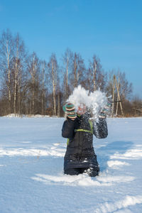 Woman throwing snow while kneeling on land against trees and sky