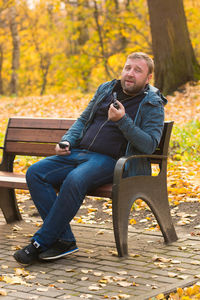 Portrait of man smoking electronic cigarette while sitting on bench against tree