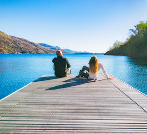 Rear view of people sitting on pier over lake against sky