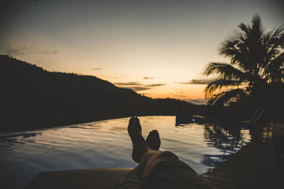Low section of man relaxing by infinity pool during sunset