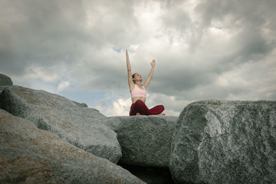 Sporty woman sitting crossed legged on rocks meditating and practicing yoga, dramatic sky.