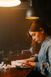 Side view of focused female worker using calculator to count income while sitting at counter with documents during work in cafe