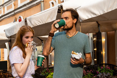 Young couple eating takeaway food and drinking coffee while standing near street cafe