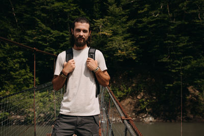 Portrait of man with backpack standing on footbridge against forest