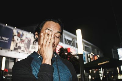 Man with hand covering face standing against built structure at night